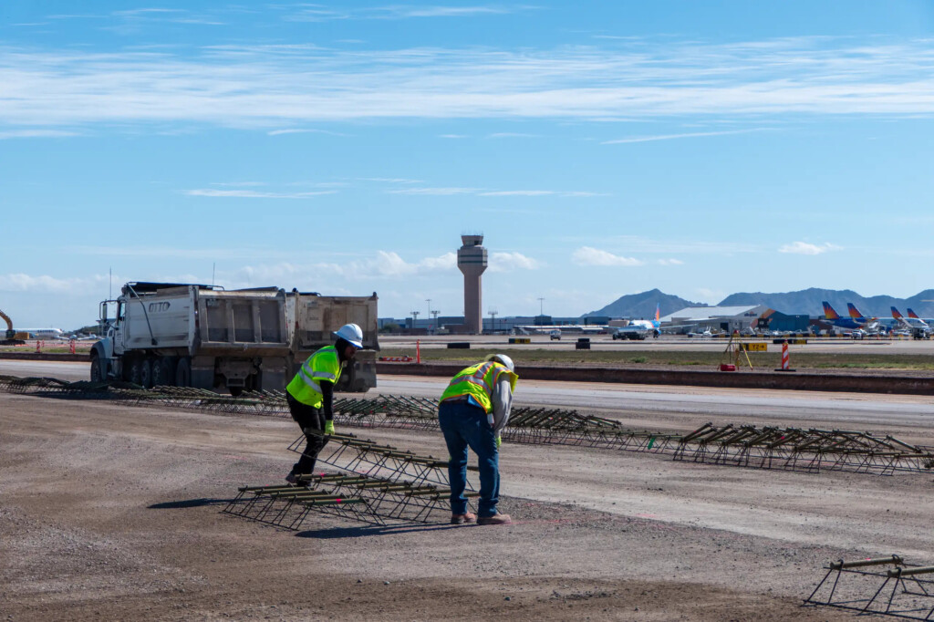 Crews in safety vests working on airfield pavement, with the air traffic control tower behind them